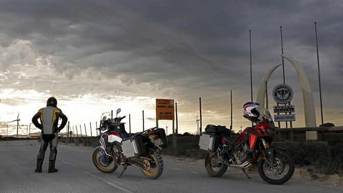 Rijder die uitkijkt op de horizon, staand naast twee Africa Twins geparkeerd op het asfalt.